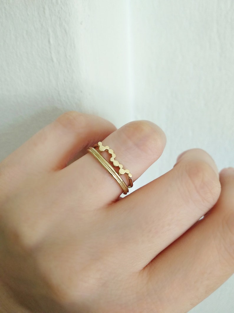 May include: Two gold rings on a finger. The top ring has a wavy design and the bottom ring is a simple band.