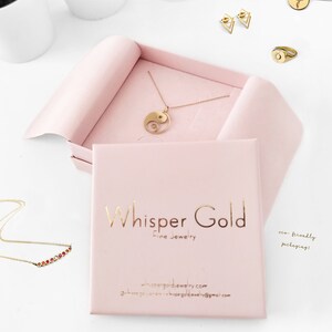 May include: A collection of gold-colored jewelry, including necklaces, earrings, and a ring, displayed on a white surface. A pink box with the text "Whisper Gold Fine Jewelry" is open, showcasing a gold Yin Yang pendant necklace. The image also includes the text "eco-friendly packaging!"