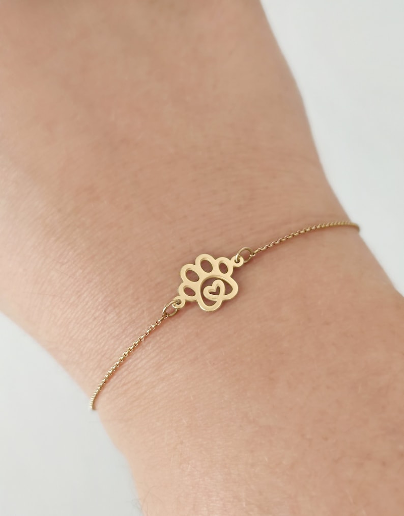 May include: A gold chain bracelet with a paw print charm featuring a heart inside. The bracelet is on a person's wrist.