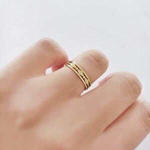 May include: A gold-colored ring with a repeating pattern of small, stylized leaf shapes. The ring is worn on a finger against a soft, neutral background. The ring's design is visible, showcasing its intricate details.