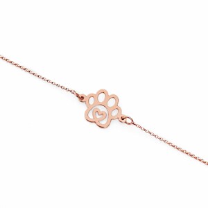 May include: A delicate rose gold chain bracelet with a paw print charm featuring a small heart inside. The charm is a cutout design, allowing light to shine through.