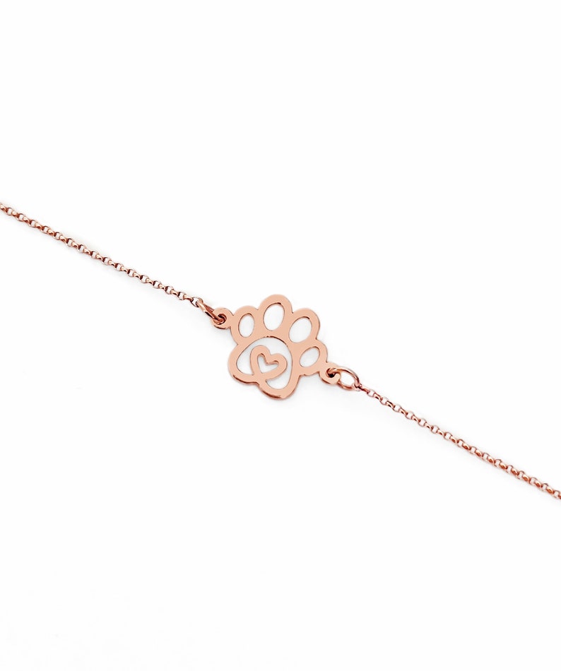 May include: A delicate rose gold chain bracelet with a paw print charm featuring a small heart inside. The charm is a cutout design, allowing light to shine through.