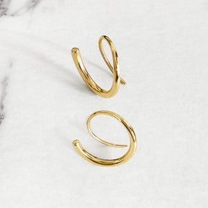 May include: A pair of gold hoop earrings. The earrings are made of a single, continuous piece of wire that is bent into a circle. The wire is slightly thicker on one side of the circle, giving the earrings a unique look.