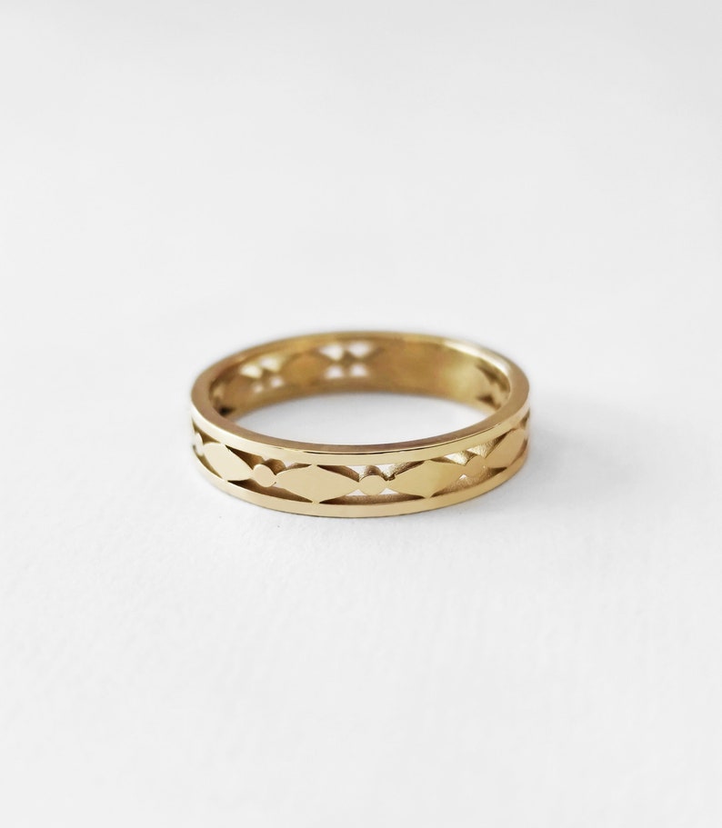 May include: A gold-colored ring with a repeating pattern of leaf-shaped cutouts and small circles. The ring is set against a white background, highlighting its intricate design and warm metallic hue. The ring is a band style.