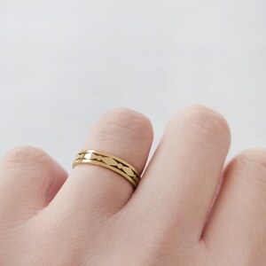 May include: A gold-colored ring with a repeating pattern of elongated ovals and small dots. The ring is worn on a finger against a white background. The ring is a simple band with a decorative design.