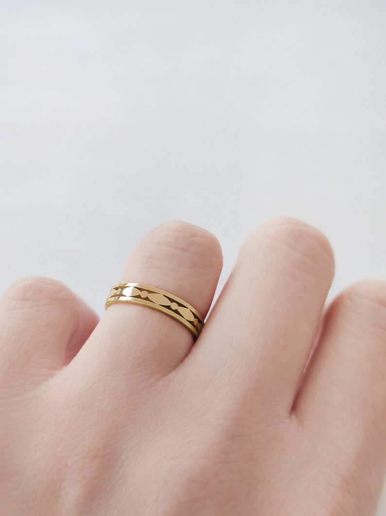 May include: A gold-colored ring with a repeating pattern of elongated ovals and small dots. The ring is worn on a finger against a white background. The ring is a simple band with a decorative design.
