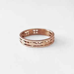 May include: A rose gold ring with a repeating pattern of cut-out shapes. The design features a series of elongated ovals and small circles, creating an openwork effect. The ring is set against a white background, highlighting its intricate details.