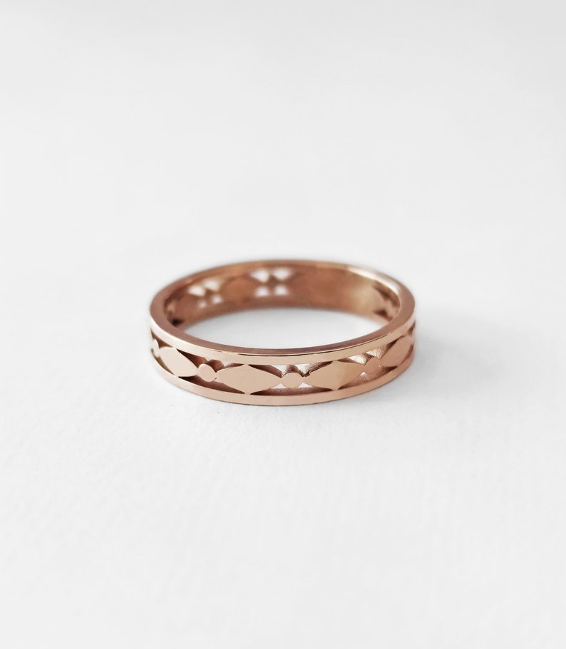 May include: A rose gold ring with a repeating pattern of cut-out shapes. The design features a series of elongated ovals and small circles, creating an openwork effect. The ring is set against a white background, highlighting its intricate details.
