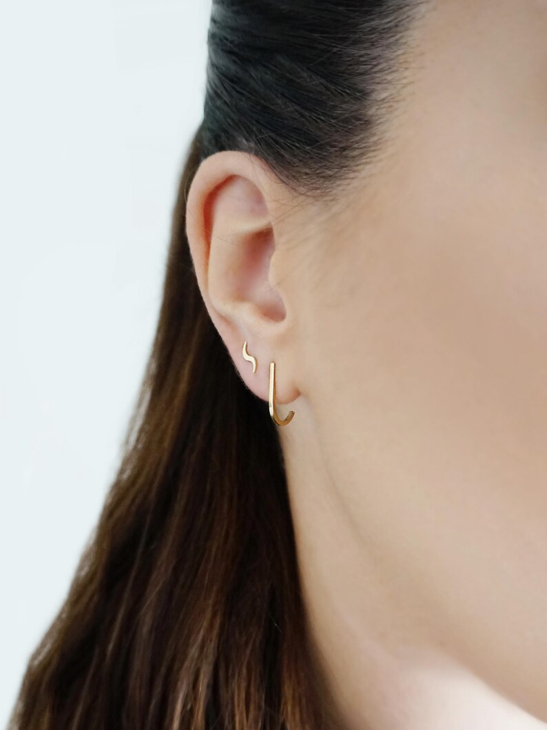 May include: A close-up of a person's ear with two gold earrings. The larger earring is a simple hoop, while the smaller earring is a curved shape.