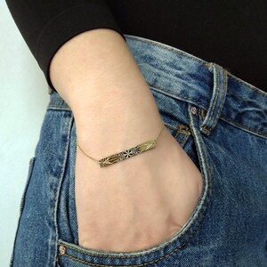 Op de afbeelding: Een gouden armband in de vorm van een staaf met een delicaat, ingewikkeld ontwerp. De armband wordt om de pols gedragen, met de staaf horizontaal gepositioneerd.