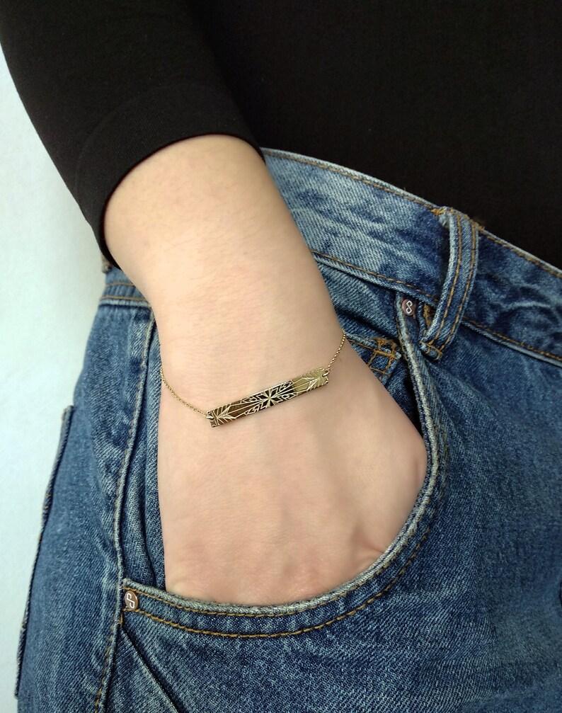 Op de afbeelding: Een gouden armband in de vorm van een staaf met een delicaat, ingewikkeld ontwerp. De armband wordt om de pols gedragen, met de staaf horizontaal gepositioneerd.