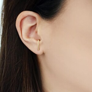 May include: A close-up of a person's ear with a gold ear cuff. The ear cuff is a thin, curved piece of metal that goes through the ear and then curves back up to the top of the ear.