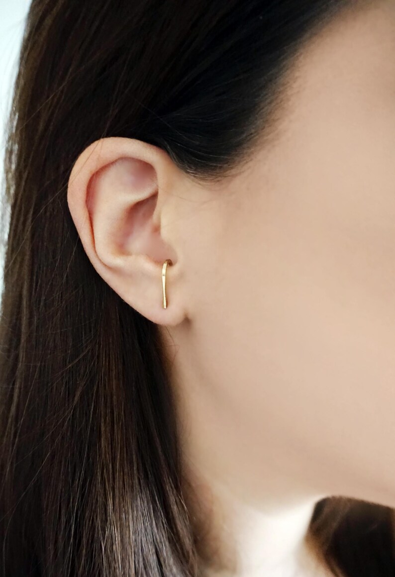 May include: A close-up of a person's ear with a gold ear cuff. The ear cuff is a thin, curved piece of metal that goes through the ear and then curves back up to the top of the ear.