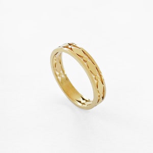 May include: A gold-colored ring with a repeating leaf-like pattern around the band. The ring is set against a white background, highlighting its intricate design and polished finish. The ring's design features a series of elongated diamond shapes.