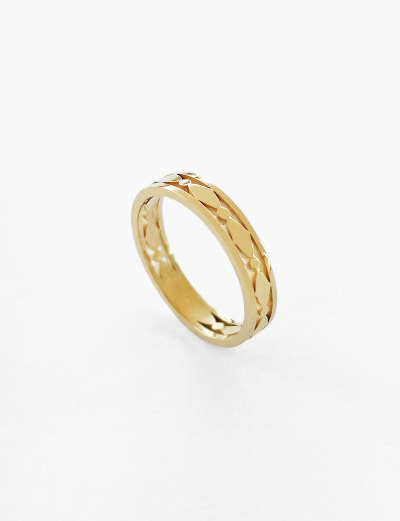May include: A gold-colored ring with a repeating leaf-like pattern around the band. The ring is set against a white background, highlighting its intricate design and polished finish. The ring's design features a series of elongated diamond shapes.