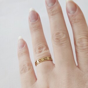 May include: A gold-colored ring with a repeating pattern of elongated shapes. The ring is worn on a finger, showcasing its design and metallic sheen. The ring's design is visible against the skin and a white background.