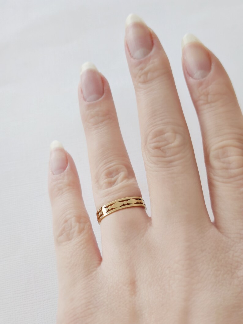 May include: A gold-colored ring with a repeating pattern of elongated shapes. The ring is worn on a finger, showcasing its design and metallic sheen. The ring's design is visible against the skin and a white background.