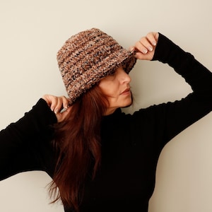 Könnte beinhalten: Ein gehäkelter Bucket Hat in Braun und Schwarz. Der Hut hat ein strukturiertes Aussehen und eine leicht gebogene Krempe. Der Hut wird von einer Person getragen, die ein schwarzes Langarmshirt trägt.