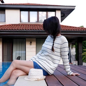 Könnte beinhalten: Eine Frau in einem weiß-blau gestreiften Pullover sitzt auf einem Holzdeck am Schwimmbad. Sie trägt einen weißen Hut mit Spitzenbesatz und eine Jeans-Shorts.