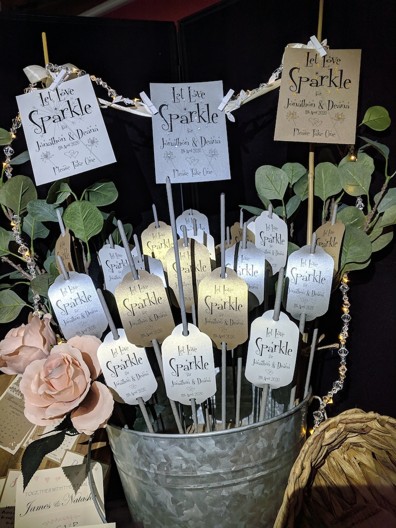 May include: A silver bucket filled with sparklers, each with a tag that reads "Let Love Sparkle for Jonathon & Deana 18 April 2020" in black script. The tags are in silver and gold with a heart design. The sparklers are surrounded by greenery and pink roses.