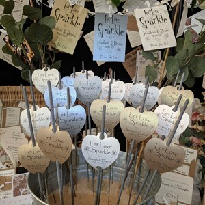 May include: A silver bucket filled with sand and heart-shaped sparklers. The sparklers have labels that say "Let Love Sparkle" with the names "Sarah & David" and the date "2nd July 2020".