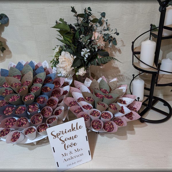 Può includere: Un tavolo con bomboniere per la cerimonia di matrimonio con due piatti bianchi pieni di piccoli coni di carta pieni di petali di rosa rosa. Un cartello recita "Sprinkle Some Love Mr & Mrs Anderson 20th July 2023".