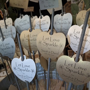 May include: A close-up of several heart-shaped wedding favor sparklers with the text "Let Love Sparkle" and the names "Sarah & David" printed on them. The sparklers are in a bucket and are ready to be given out to guests.