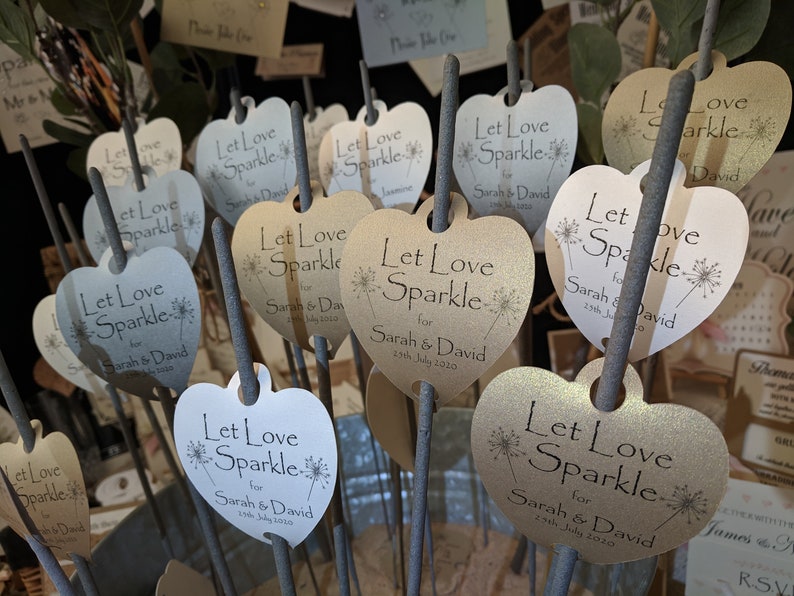 May include: A close-up of several heart-shaped wedding favor sparklers with the text "Let Love Sparkle" and the names "Sarah & David" printed on them. The sparklers are in a bucket and are ready to be given out to guests.