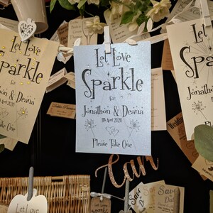May include: A wedding favor table with a variety of cards that say "Let Love Sparkle" with the couple's names and wedding date. The cards are decorated with glitter and small white flowers.