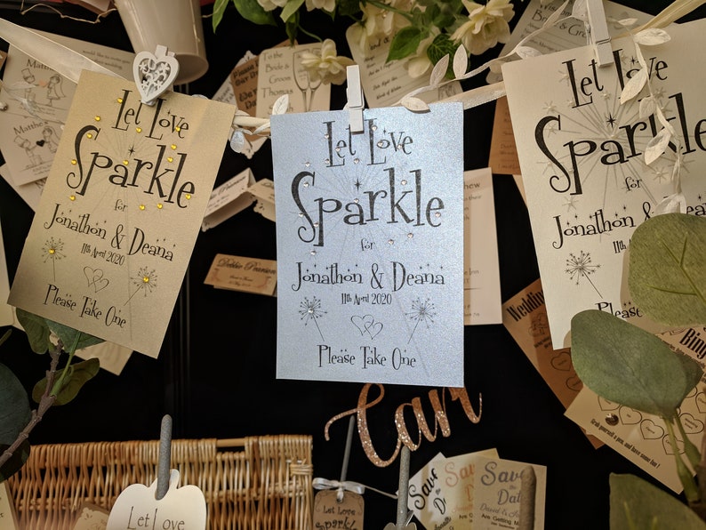 May include: A wedding favor table with a variety of cards that say "Let Love Sparkle" with the couple's names and wedding date. The cards are decorated with glitter and small white flowers.