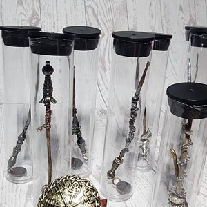 May include: Collection of decorative wands in clear tubes with black lids. The wands have intricate designs and are various colors. A gold-colored sphere with a detailed pattern sits on a wooden stand in the foreground.