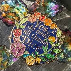 Op de afbeelding: Een gouden emaille pin met een blauwe achtergrond en de tekst "Ja, ik heb al deze dobbelstenen nodig". De pin is omringd door kleurrijke, iriserende dobbelstenen.