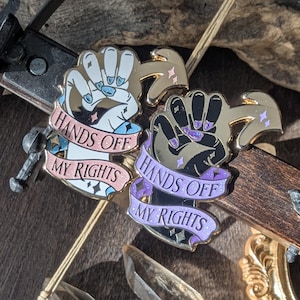 May include: Two enamel pins with the text "HANDS OFF MY RIGHTS" and a fist design, one with a light skin tone and the other with a dark skin tone. A small crossbow, two clear quartz crystals, and a gold-colored decorative frame are also visible.