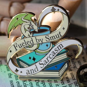 May include: Enamel pin with a dragon perched on a coffee cup, above a stack of books. The pin has a banner that reads "Fueled by Smut and Sarcasm." The pin is white, teal, and gold.