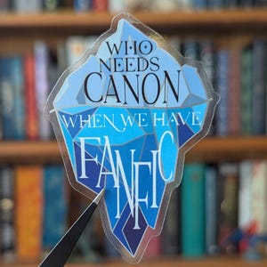 Puede incluir: Una pegatina transparente con el texto "WHO NEEDS CANON WHEN WE HAVE FANFIC" en azul y blanco, con forma de iceberg. La pegatina está sujeta con unas pinzas delante de una estantería llena de libros.