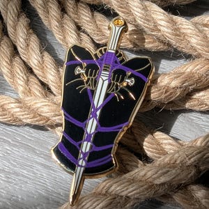 Puede incluir: Un pin de esmalte negro y dorado con un diseño de espada en morado y blanco. El pin se encuentra sobre un fondo de gruesa cuerda enrollada.