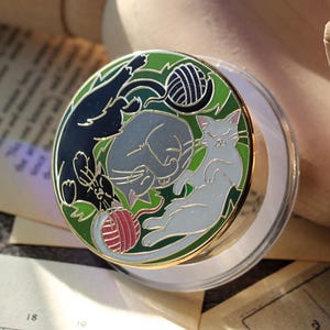 May include: A round, colorful enamel pin featuring three cats and a ball of yarn. The pin has a green, black, and white color scheme with gold accents. The cats are in various poses, with one sleeping, one playing, and one looking up.