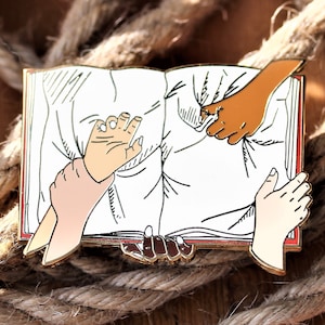 Könnte beinhalten: Goldfarbene Emaille-Anstecknadel, die drei Hände zeigt, die aus einem offenen Buch greifen. Die Hände haben unterschiedliche Hautfarben und symbolisieren Einheit und Inklusion.