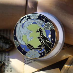 May include: A circular enamel pin depicting two dragons, one white and one black, set against a pale green backdrop. The pin has a dark blue border with stars and clouds. The pin is set on a clear, round base.