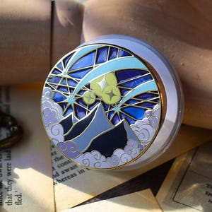 May include: A round, clear glass bottle stopper with a detailed enamel design. The design features a night sky scene with mountains, stars, and clouds in shades of blue, purple, and gold, with a gold-coloured metal rim.