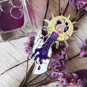 May include: A silver and gold enamel pin featuring a hand pouring purple liquid into a circular design with a flame in the center. The design is surrounded by a yellow circle with black text.