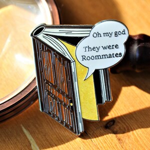 May include: Enamel pin in the shape of a book with a door design. The pin has a speech bubble with the text "Oh my god They were Roommates". The book has a yellow glittery page.