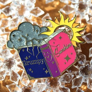 Può includere: Uno spillo in smalto dorato con due libri aperti, uno blu con la parola "Grumpy" e l'altro rosa con la parola "Sunshine". Una nuvola di glitter argentata è sopra i libri e un sole dorato con raggi è sopra il libro rosa.
