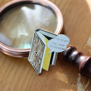May include: A silver enamel pin in the shape of an open book with yellow pages and a speech bubble that says "Oh my god They were Roommates". A copper-colored magnifying glass is in the background.