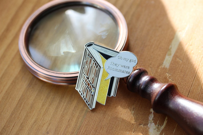 May include: A silver enamel pin in the shape of an open book with yellow pages and a speech bubble that says "Oh my god They were Roommates". A copper-colored magnifying glass is in the background.