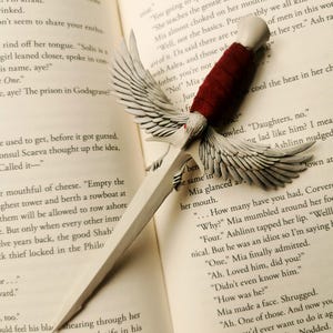 May include: A white sword bookmark with a red handle and white wings. The bookmark is resting on an open book with text about a prison in Godsgrave.