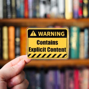 Könnte beinhalten: Ein rechteckiger Aufkleber mit gelbem Hintergrund und schwarzem Rand. Der Aufkleber trägt die Aufschrift "WARNING Contains Explicit Content" in fetter schwarzer Schrift. Oben befindet sich ein schwarz-gelbes Gefahrensymbol. Der Aufkleber wird vor einem Bücherregal gehalten.