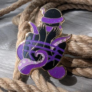 May include: A black and purple enamel pin depicting a stylized figure bound with purple ropes and tentacles. The figure wears a black hat and blindfold. The pin has a gold border and is set against a background of thick, brown rope.