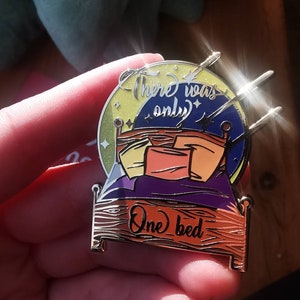 Puede incluir: Un pin de esmalte dorado con un dise&ntilde;o de cama con cabecera morada y almohadas de colores. La cama est&aacute; rodeada de una luna amarilla con estrellas y el texto "There was only one bed".