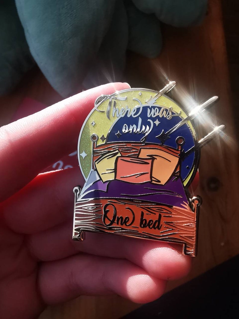 Puede incluir: Un pin de esmalte dorado con un dise&ntilde;o de cama con cabecera morada y almohadas de colores. La cama est&aacute; rodeada de una luna amarilla con estrellas y el texto "There was only one bed".